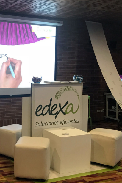 Stand publicitario de la marca Edexa con el eslogan Soluciones eficientes, acompañado de banquetas blancas y un telón de proyección al fondo.