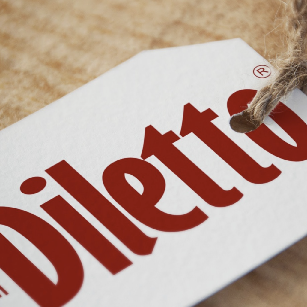 Plano detalle en primer plano de una etiqueta colgante de color blanco con la marca Diletto impresa en letras rojas, sujeta por una cuerda de yute artesanal.