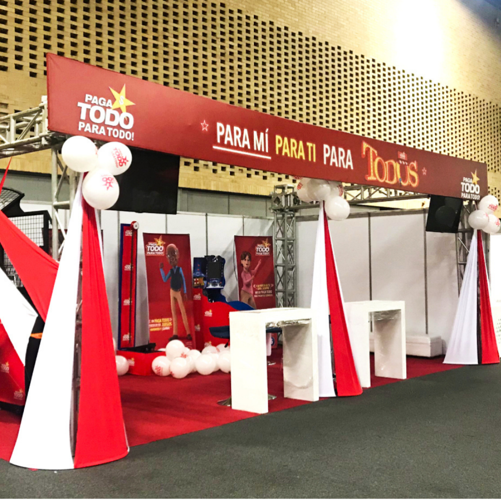 Gran stand publicitario de la marca Paga Todo en una feria, con letreros rojos que muestran el eslogan Para mí para ti para todos, mesas altas blancas y decoración con globos.